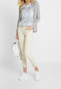 Pantaloni slim-fit beige chiaro, cardigan grigio a trama, blusa blu con fantasia, sneakers bianche e borsa bianca. Cuffie dei pantaloni arrotolate visibili.