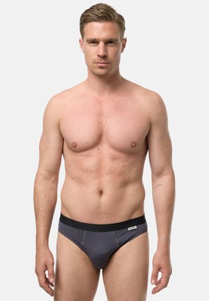 Wysportowany dorosły mężczyzna z krótkimi włosami, ubrany w ciemnoszare majtki z czarnym pasem, stojący na jednolitym białym tle.