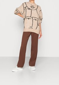 Beige tröja med svarta abstrakta ansiktsmönster, matchad med bruna raka byxor och vita sneakers. Mjuk stickad textur synlig.