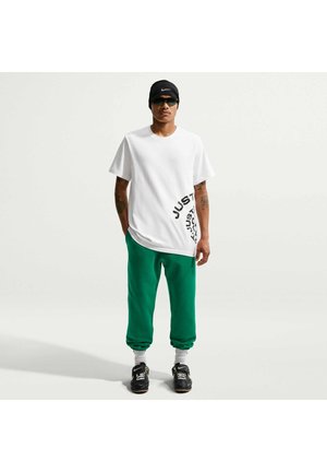 Uomo che indossa una t-shirt bianca con grafica, pantaloni della tuta verdi, sneakers nere, fascia nera e occhiali da sole, in piedi davanti a uno sfondo chiaro uniforme.