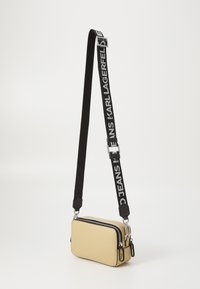 Béžová obdĺžniková crossbody taška s textúrovaným povrchom, čierny nastaviteľný popruh s bielym písmom a striebornými zipsovými kovaniami.