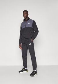 Puma Trainingsanzug in Schwarz und Grau. Jacke mit Reißverschluss und hohem Kragen; die Hose hat elastische Bündchen. Sportschuhe in Schwarz mit olivgrünen Akzenten.