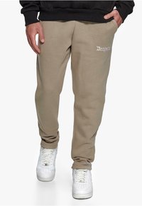 Beige sweatpants in cotton blend, featuring an embroidered logo on the left thigh, relaxed fit, and cuffed ankles paired with white sneakers.