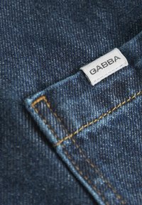Tamno plavi traper tkanina s vidljivim dizajnom džepa i narančastim šavovima; bijela oznaka s natpisom "GABBA" nalazi se blizu šava.