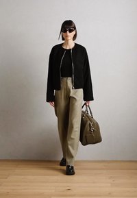 Černá texturovaná bomber bunda, černé tričko, béžové kalhoty s vysokým pasem, černé boty, držící olivově zelenou tašku, s tmavými slunečními brýlemi.