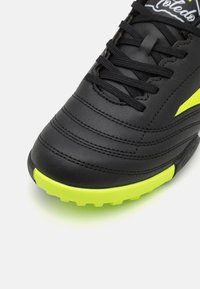 Botas de fútbol negras con parte superior de cuero texturizado, que presentan acentos amarillos en el costado y una suela de goma amarilla brillante con tacos.