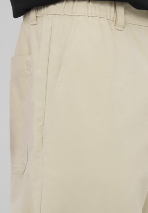 Pantalon beige avec taille élastique, poche latérale avec rabat, et détails de couture visibles à l'avant et sur les côtés.