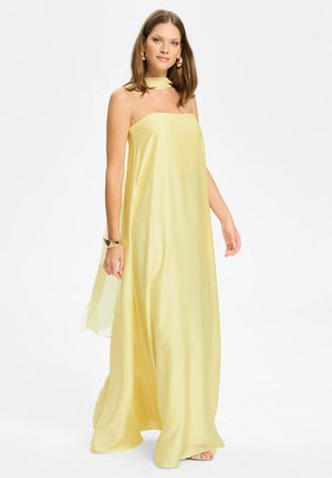 Nainen seisoo pukeutuneena olkaimettomaan, liehuvaan vaaleankeltaiseen mekkoon, jolla on yhtenevä kaulahuivi ja oikeassa ranteessaan kultaista rannekorua.