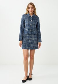 Kobieta w niebiesko-białej w kratę kurtce z złotymi guzikami i dopasowanej spódnicy, w czarnych balerinach na kostkę, stojąca na białym tle.