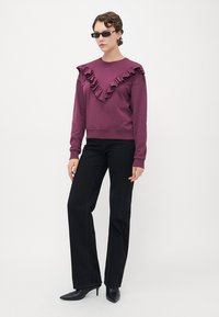Persona che indossa un top viola con ruches, pantaloni neri a gamba dritta, scarpe con tacco a punta nere e occhiali da sole rettangolari piccoli.