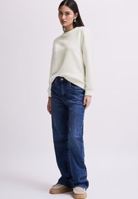 Maglione verde chiaro a trama con motivo a zigzag, abbinato a jeans dritti blu scuro e scarpe beige senza lacci.