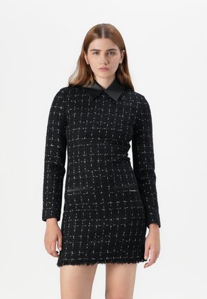 Jeune femme avec de longs cheveux portant une robe noire ajustée avec un motif texturé à carreaux et un col noir, faisant face à l'avant sur fond blanc.