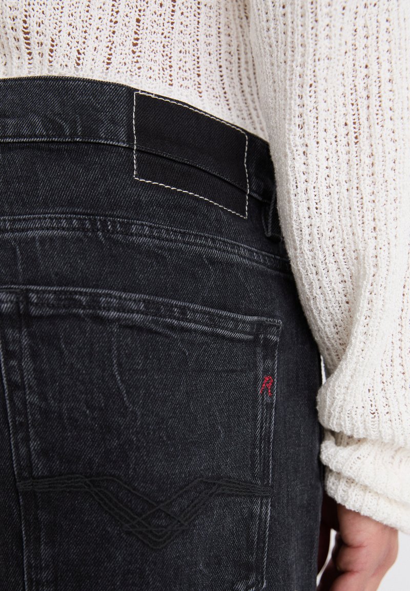 Schwarze Jeans aus Denim mit einer subtilen Webtextur, ausgestattet mit weißem Nähen und rotem Branding auf der Gesäßtasche. Sichtbares Detail am Taillenbund.