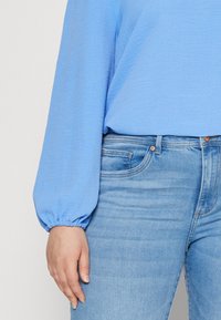 Blusa azzurra chiara, con texture e maniche lunghe arricciate, abbinata a jeans in denim azzurro chiaro con tasca frontale e dettagli con bottone.