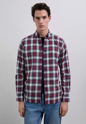 Joven vestido con una camisa de cuadros en rojo, blanco y verde sobre una camiseta negra, con jeans azules, de pie contra un fondo claro y liso.