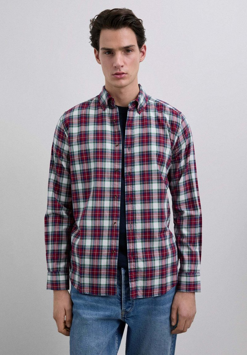 Giovane uomo che indossa una camicia a quadri rossa, bianca e verde sopra una maglietta nera con jeans blu, in piedi contro uno sfondo chiaro uniforme.