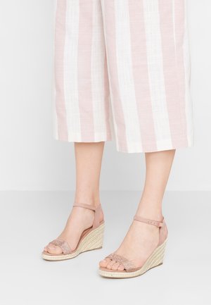 Kobieta nosząca różowe i beżowe sandały na klinie z paskami wokół kostek oraz szerokie, prążkowane spodnie o długości do kostek.