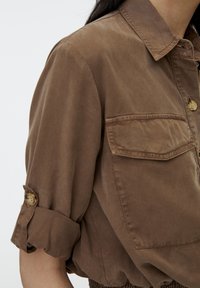 Chemise brune boutonnée avec manches retroussées et poche poitrine sur une personne devant un fond uni.