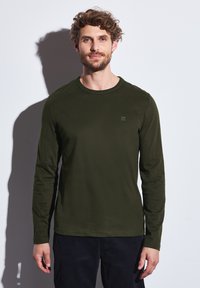 Camisa de manga larga verde oscuro con un escote redondo. Presenta un pequeño y sutil logo en el pecho. Tejido suave con un diseño ajustado.