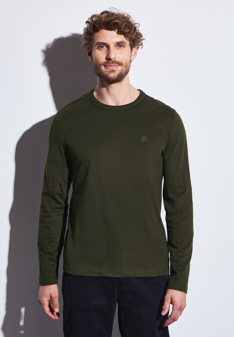 Camisa de manga larga verde oscuro con un escote redondo. Presenta un pequeño y sutil logo en el pecho. Tejido suave con un diseño ajustado.