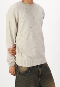 Personne portant un pull beige en tricot avec des motifs en losange et un jean noir délavé, tenant son bras gauche derrière le dos.