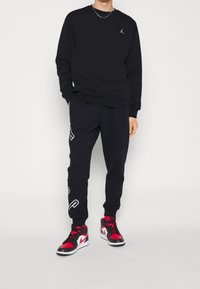 Sudadera negra con un pequeño logo, pantalones jogger negros a juego con un diseño blanco, y zapatillas rojas y negras con detalles en blanco.