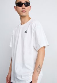 Tricou oversized din bumbac alb, cu un mic logo Adidas negru pe piept. Purtați cu ochelari de soare negri eleganți, evidențiind un stil minimalist.
