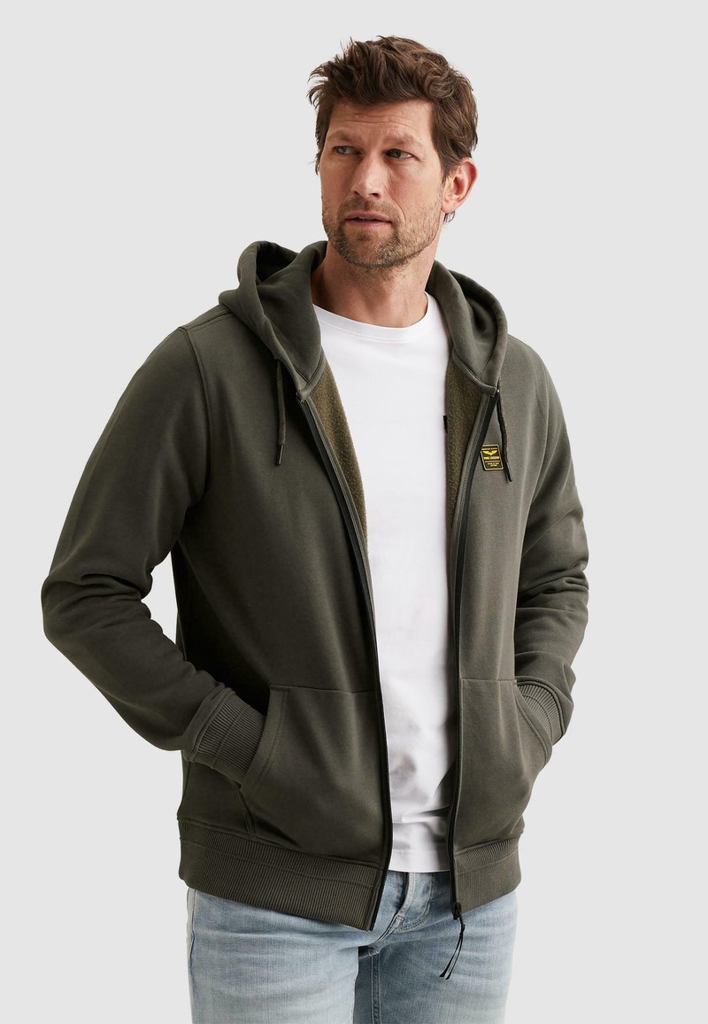 Homme portant un sweat à capuche zippé vert olive sur un t-shirt blanc et un jean bleu clair, les mains dans les poches, regardant sur le côté.