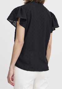 Schwarze Kurzarmbluse mit strukturiertem Polka-Dot-Muster, leicht gerafften Ärmeln und abgerundetem Saum. Der Stoff wirkt leicht und weich.
