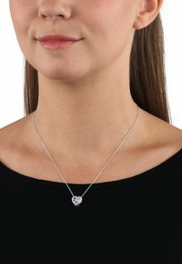 Collana d'argento con ciondolo a forma di cuore che presenta una gemma viola, esposta su un collo liscio e nudo contro un top nero.