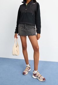 Person i svart fleecetröja och grå jeanskjol som håller en beige handväska, bär beige och vinröda sneakers på blått golv.