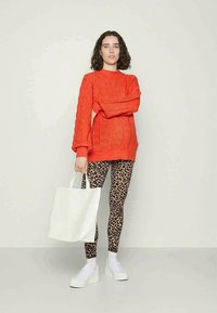 Pull en tricot orange vif avec un motif en relief, associé à un legging imprimé léopard marron et des baskets blanches ; tenant un sac à dos blanc.