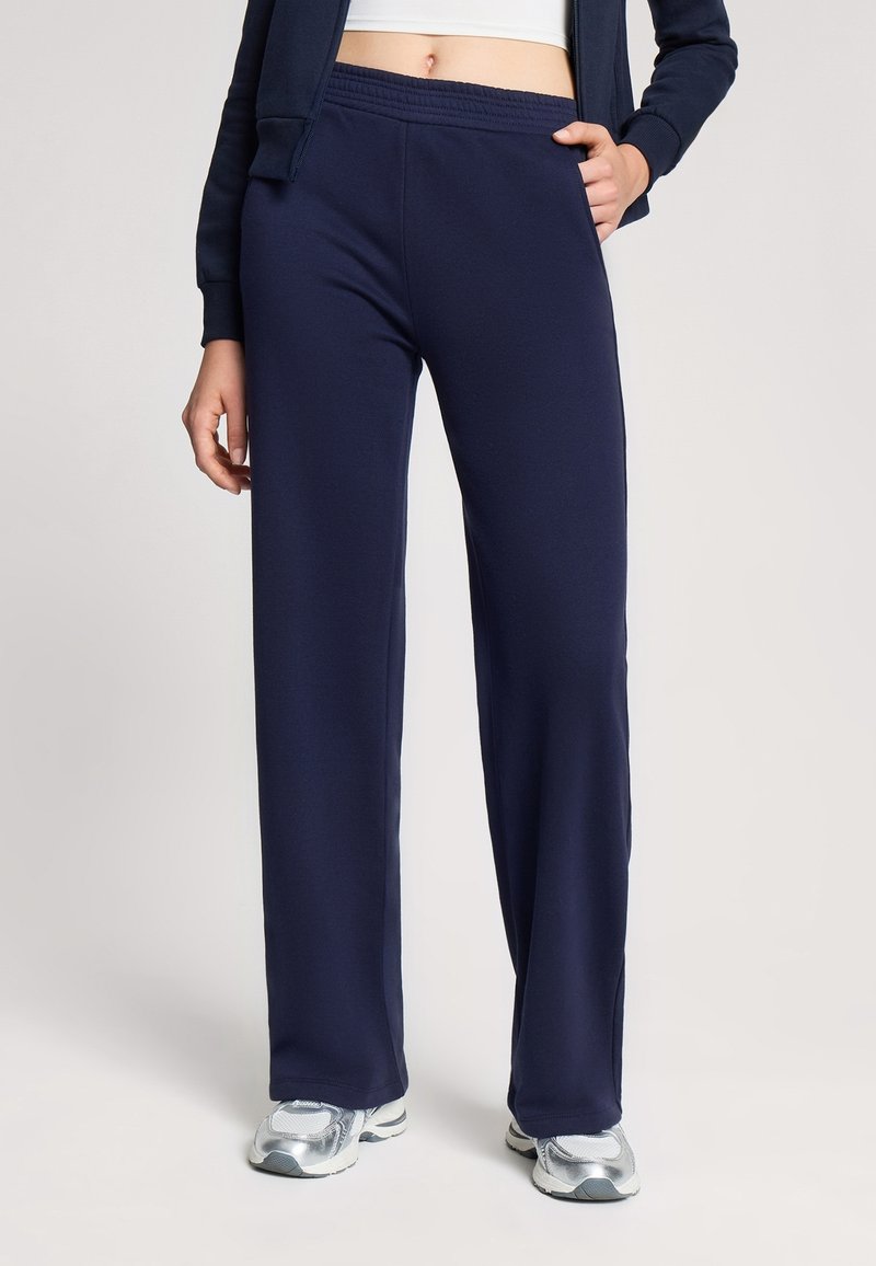 Pantaloni della tuta blu navy realizzati in tessuto morbido, con una vestibilità comoda, vita elastica e tasche laterali. Abbinati a scarpe da ginnastica argento.