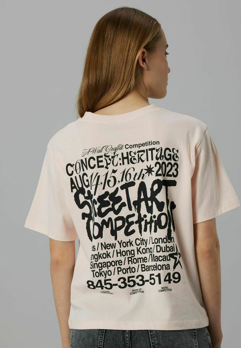 Ljusrosa t-shirt med rund hals som har fet svart text på baksidan, där det beskrivs en gatukonsttävling med olika staders namn och datum.