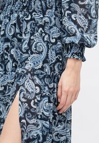 Robe à motif paisley bleu et blanc avec taille smockée et poignets de manches, avec une fente haute à la cuisse et une main visible reposant sur le côté.