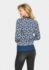 Pull en tricot avec un motif abstrait bleu et blanc, col et poignets côtelés, et un ourlet bleu uni. Associé avec un jean foncé.