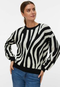 Vrouw in een zwart-witte zebra gestreepte trui en blauwe jeans, poserend met haar handen op haar heupen tegen een effen achtergrond.