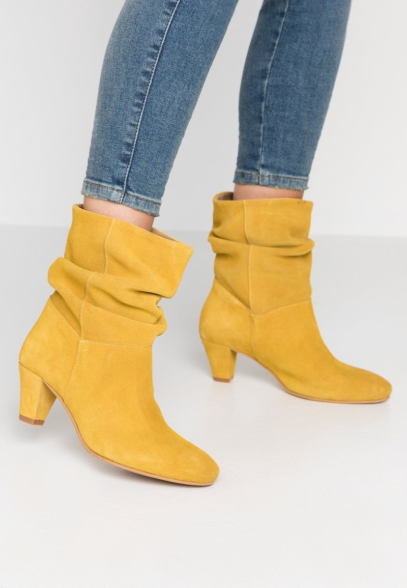 Femme portant des bottes jaunes en daim à talons souples mi-mollet associées à un jean skinny bleu sur un sol blanc.