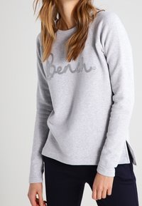 Sweat-shirt gris clair à manches raglan, avec un logo "Bench" texturé sur le devant et des fentes latérales à l'ourlet. Texture de tissu douce.