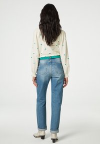 Blusa crema con motivo floreale e maniche a sbuffo abbinata a jeans dritti azzurri; il modello indossa una cintura verde e stivaletti.