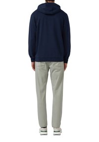 Uomo che indossa un cappuccio blu navy, pantaloni grigi chiari e sneakers bianche, in piedi con la schiena rivolta verso l'osservatore su uno sfondo bianco.