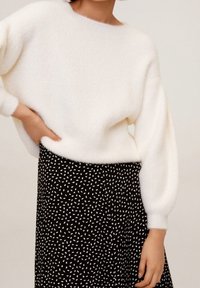 Persoon in een wit, fluffy sweater gecombineerd met een zwarte rok met een klein wit stippenpatroon, poseert met één hand op de heup.