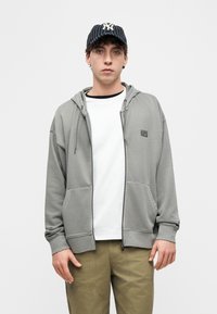 Νεαρός άνδρας που φοράει ριγέ καπέλο μπέιζμπολ, γκρι φούτερ με φερμουάρ πάνω από λευκό T-shirt και χακί παντελόνι cargo, στέκεται μπροστά σε απλό φόντο.