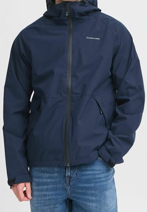 Uomo che indossa una giacca impermeabile North Bend blu navy con cappuccio e zip frontale, abbinata a jeans blu, mani nelle tasche.