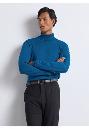Uomo con le braccia incrociate che indossa un maglione dolcevita blu e pantaloni neri, in piedi contro un muro bianco, guardando di lato.