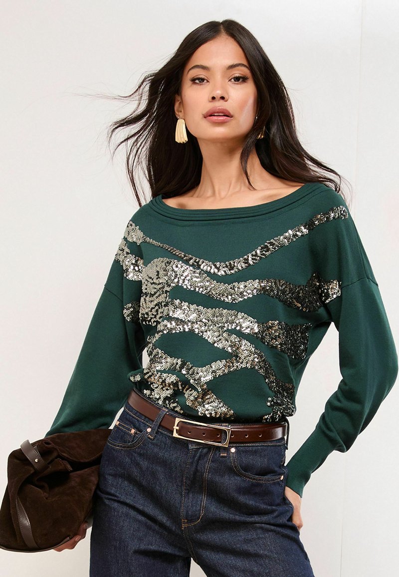 Zelený svietiaci sveter s off-the-shoulder dizajnom, s trblietavými striebornými cik-cak vzormi, skombinovaný s tmavými džínami a hnedou taškou.