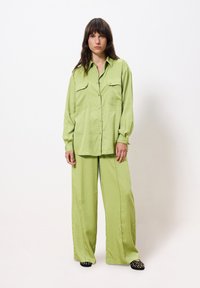 Limoengroene, langemouwenshirt met twee borstzakken en een relaxte pasvorm, gecombineerd met bijpassende broek met wijde pijpen en zwart-bruin gemusterde schoenen.