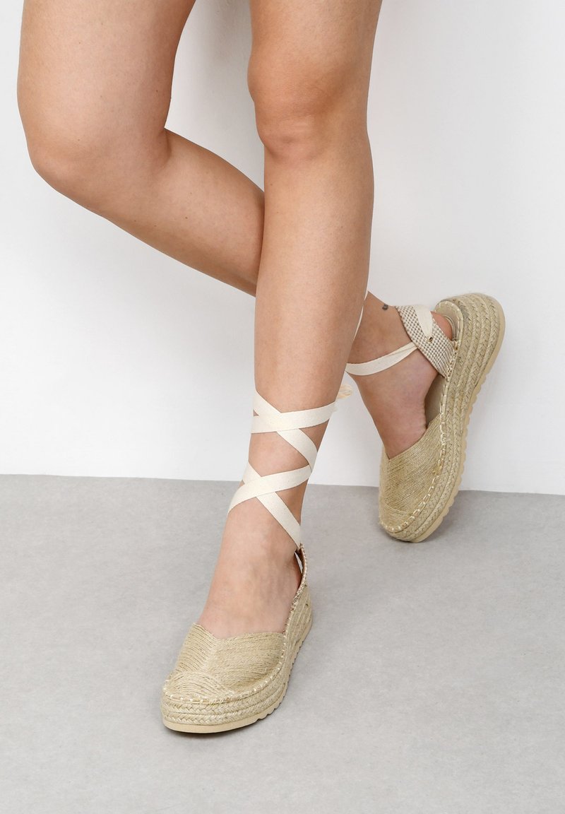 Espadrille beige con parte superior de yute tejido, suela de goma y tiras de tela entrelazadas alrededor del tobillo. Diseño de plataforma baja.