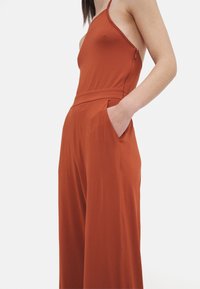 Combinaison de couleur rouille avec un haut ajusté, des bretelles spaghetti et un pantalon large. Dispose de poches latérales et d'une texture lisse et douce.