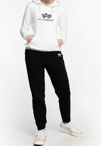 Vit hoodie med en svart logotyp, matchad med svarta sweatpants som har små logotyper. Kläderna är avslappnade och tillverkade av mjukt material.
